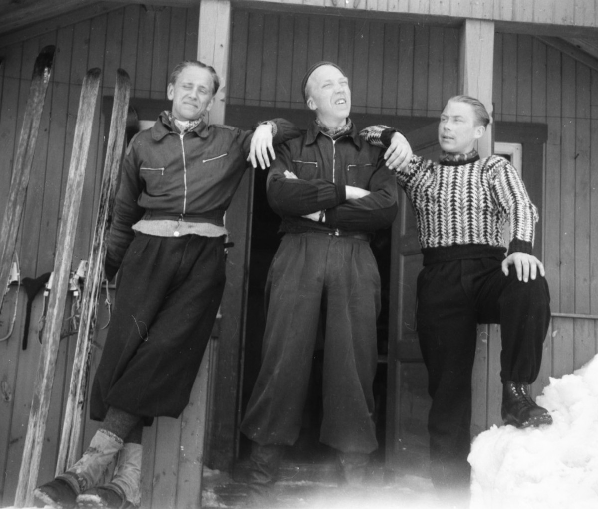 Black and white photo of three men posing in front of cabin and skis