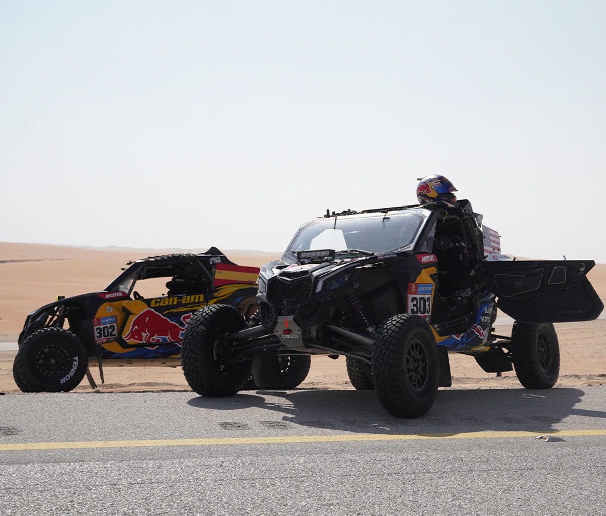 Two Dakar side by sides parked on a paved road with the desert in the background.