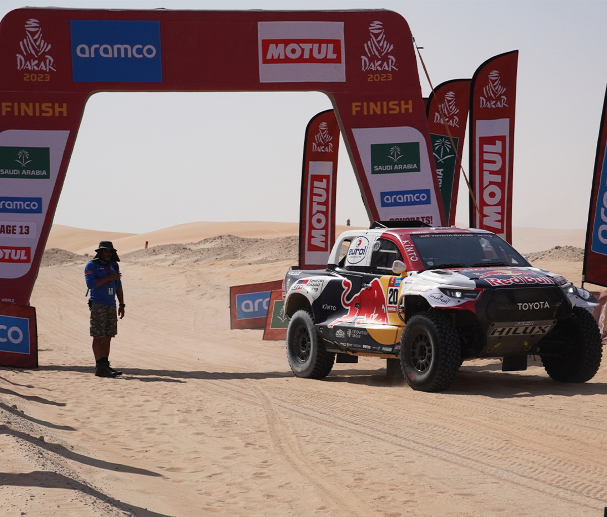 Finish line at the Dakar Rally with a man standing under an arch with a Toyota off-road truck driving through.