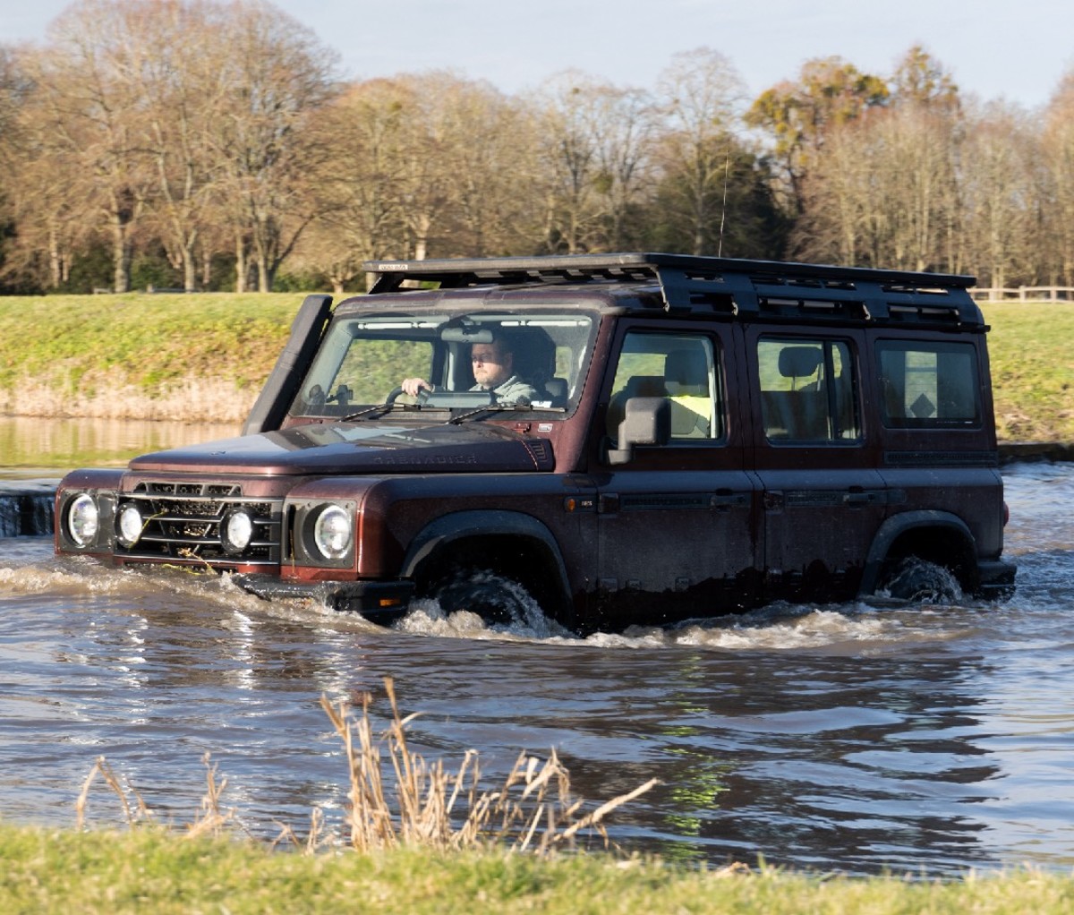 Boxy maroon 4x4 SUV driving through a deep pond with a field and trees in the background.